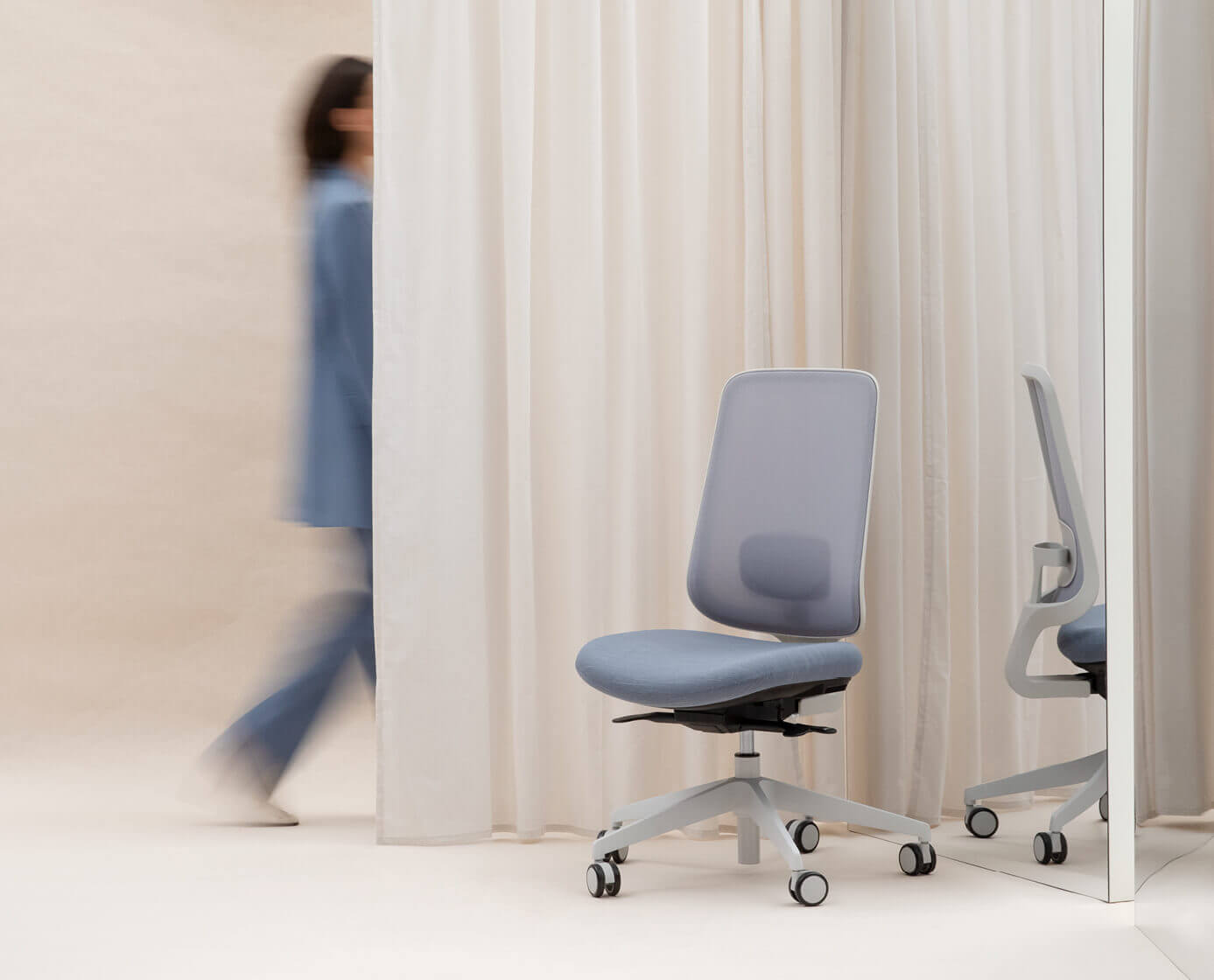 Task One Grey Frame Office Chair In Front Of A Mirror And Cream Curtain With Someone Walking Behind It