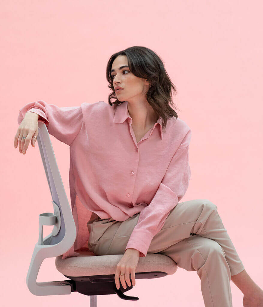 Model Wearing A Pink Shirt And Cream Trousers Rotating Around On A Task One Grey Frame Office Chair With No Arms