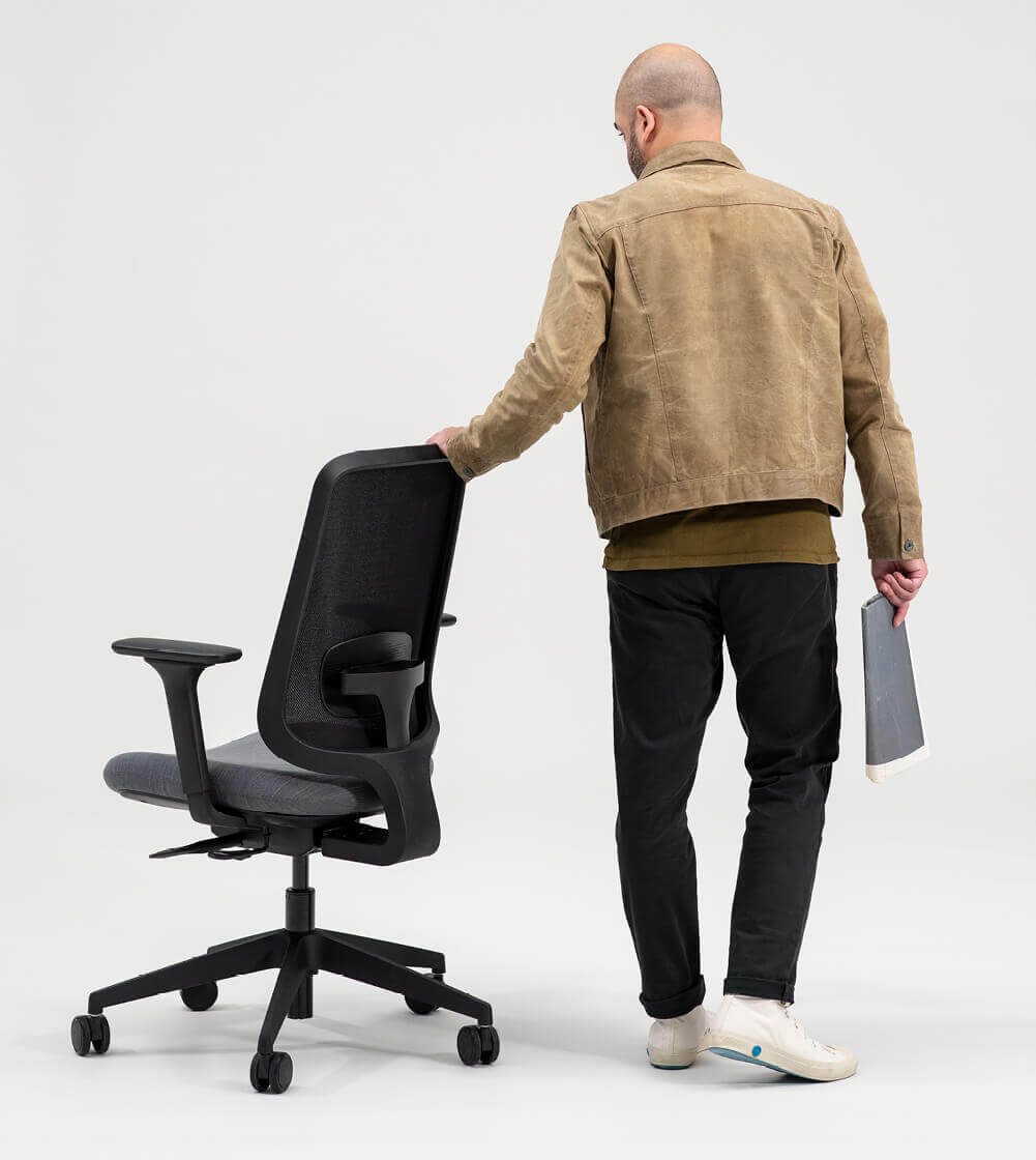 A Person Standing Behind a Task One Black Frame Office Chair and Holding It With Their Left Hand