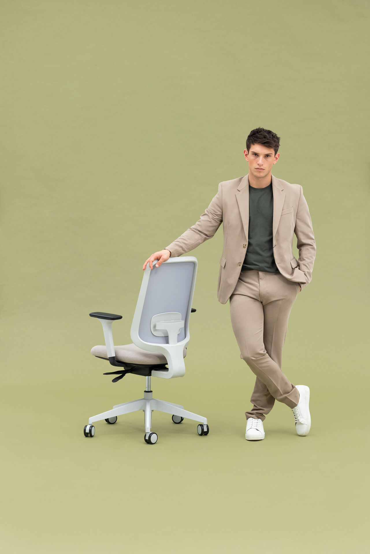 Person Facing You With One Leg In Front Of The Other Holding Onto The Back Of A Task One Grey Frame Office Chair With Arms