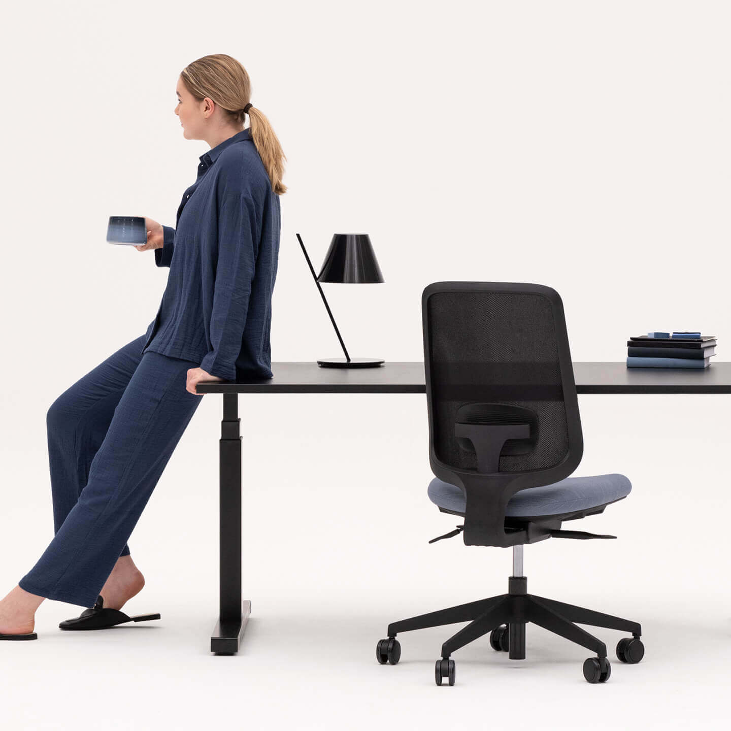 Person Holding a Mug Leaning Against a Height Adjustable Black Frame Desk With Black Top