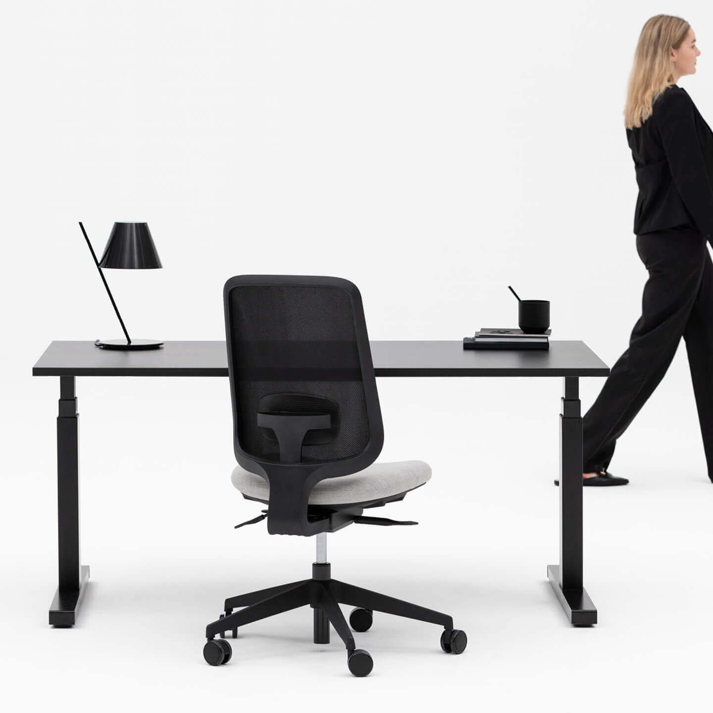 Person Walking By a Height Adjustable Black Desk and Black Framed Office Chair With No Arms