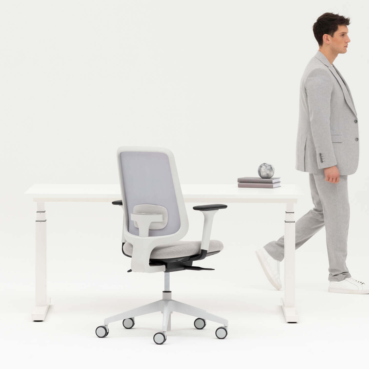 Person Walking By A White Height Adjustable Desk and Grey Framed Office Chair With Arms