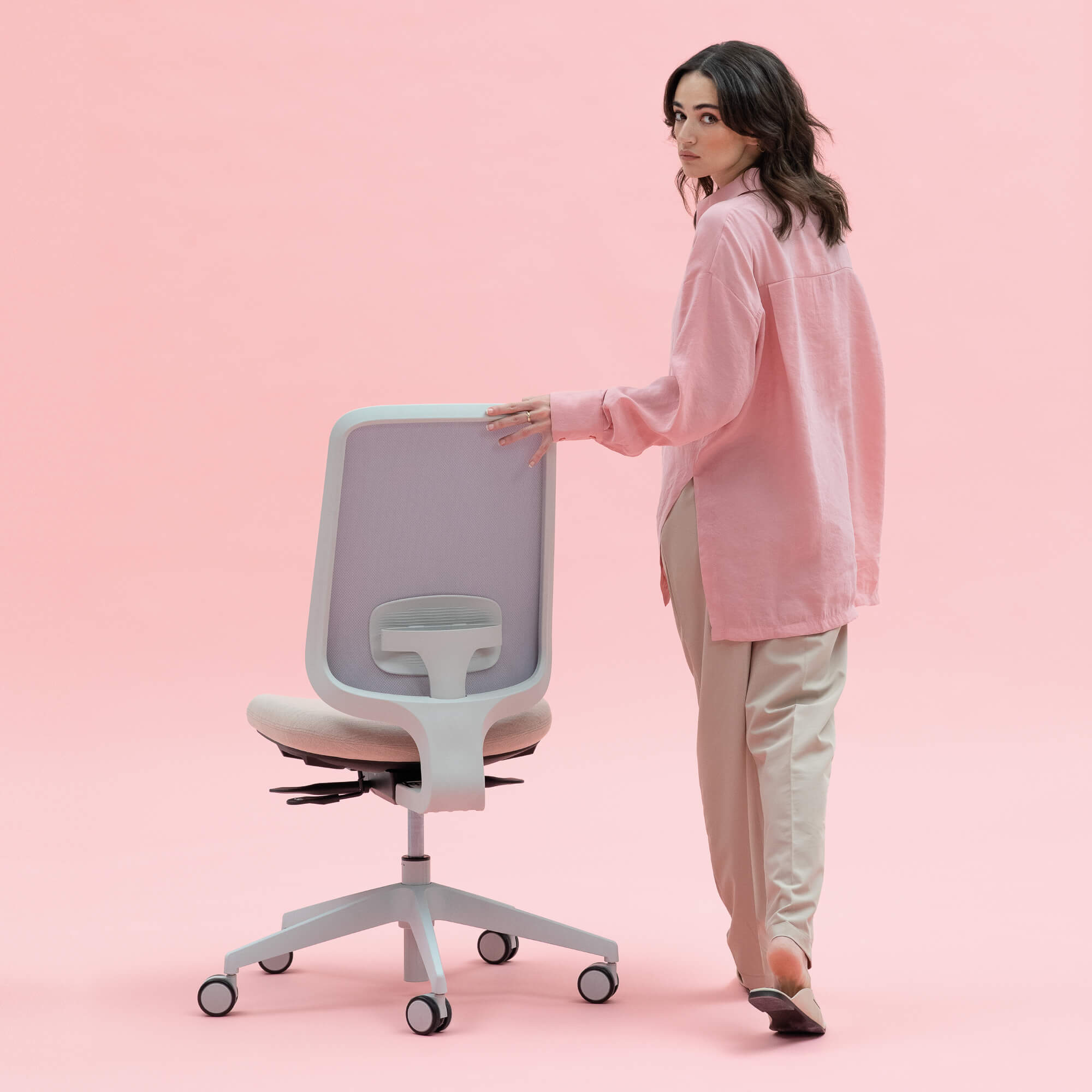 Model Wearing A Pink Shirt And Cream Trousers Turning Around Whilst Holding Onto The Back Of A Task One Office Chair