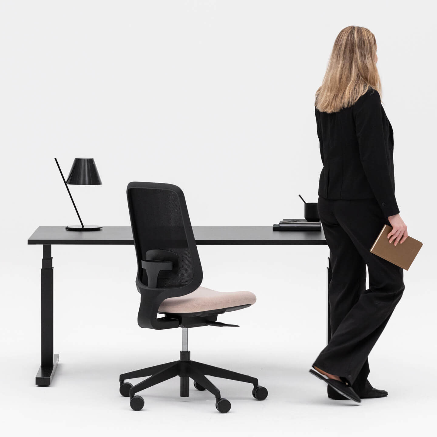 Model Wearing All Black Holding A Notepad Walking Away From A Black Task One Office Chair And Desk One Height Adjustable Desk