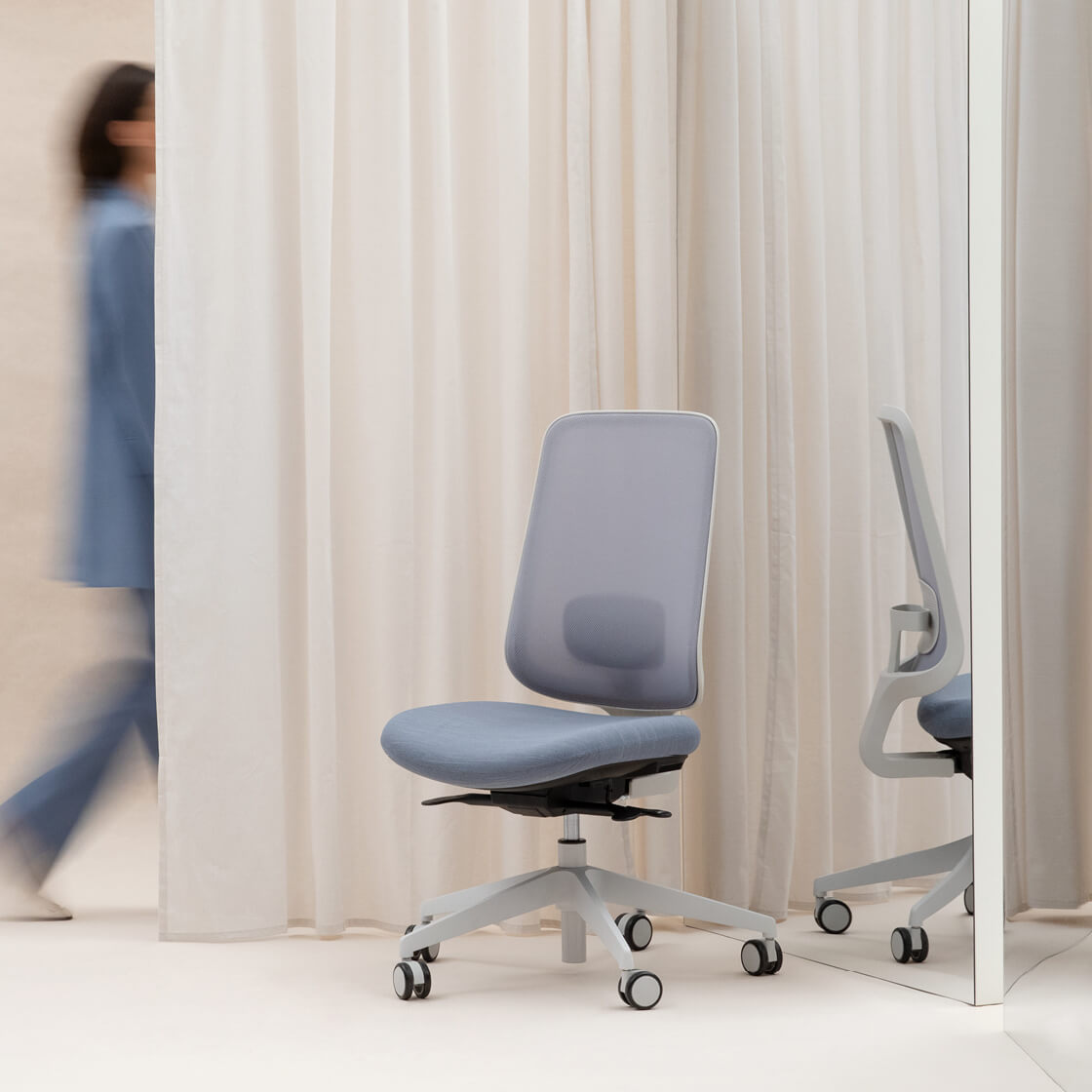 Task One Grey Frame Office Chair In Front Of A Mirror And Cream Curtain With Someone Walking Behind It