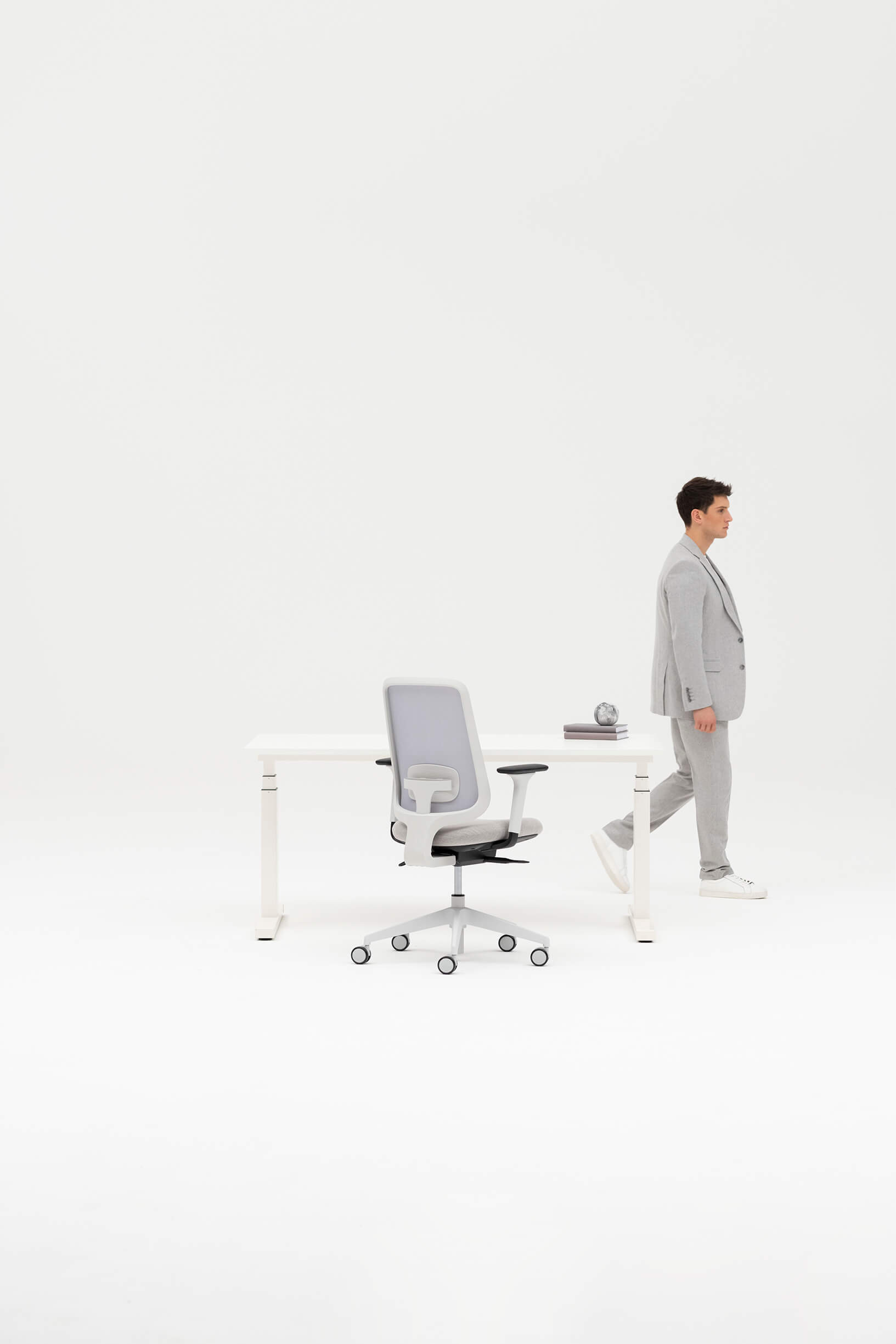 Model In Grey Trousers and Blazer Walking In Front Of A White Desk One Adjustable Height Desk And Task One Chair