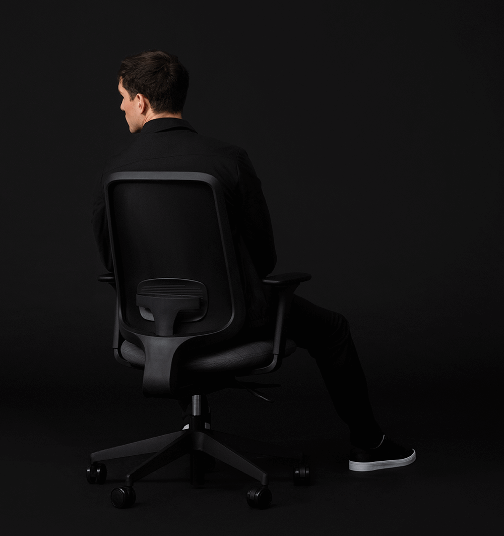 The Back Of A Model Wearing All Black Sitting On An All Black Task One Office Chair With Arms