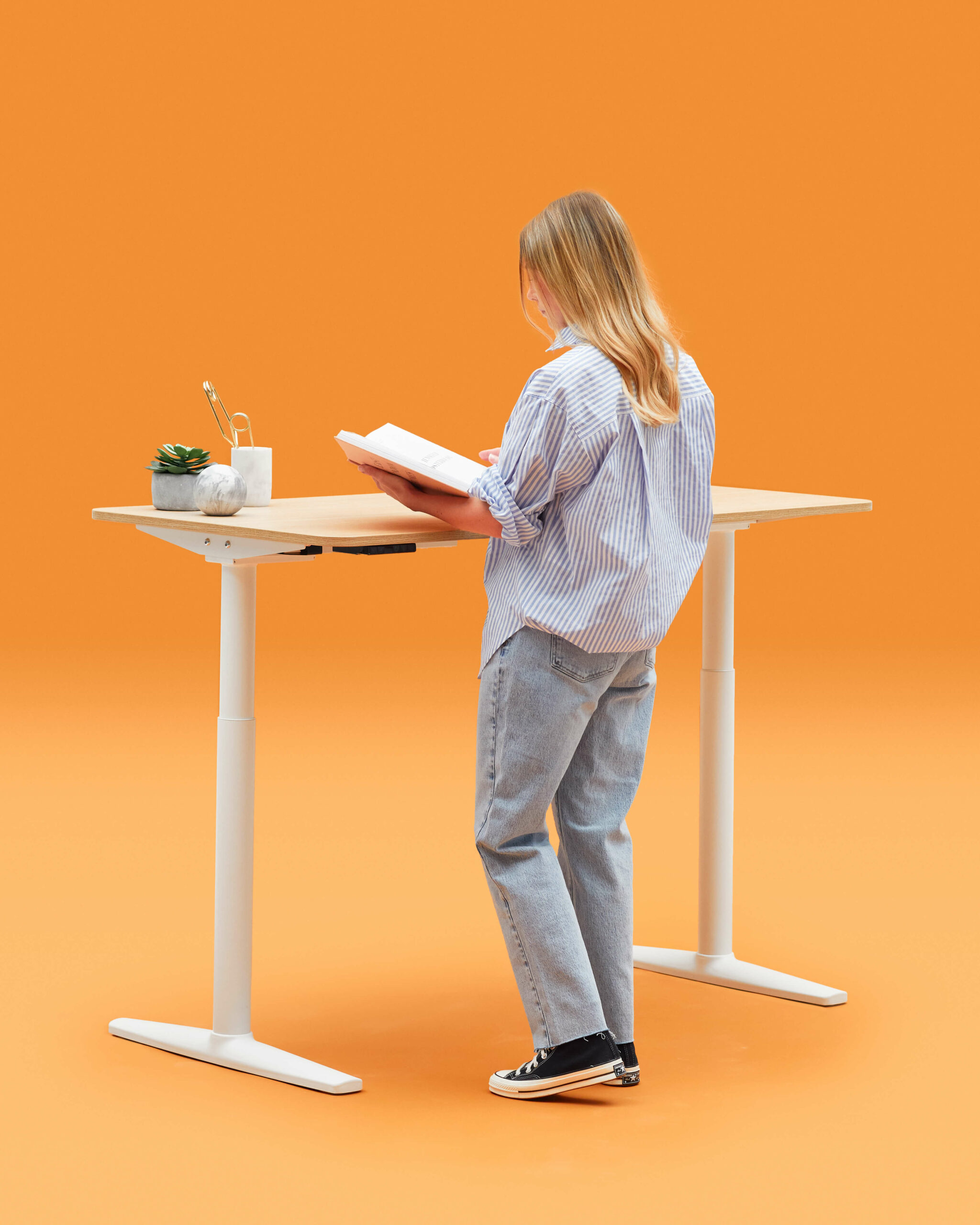 Person Reading a Book In Front Of a Height Adjustable White Frame Desk With Oak Top