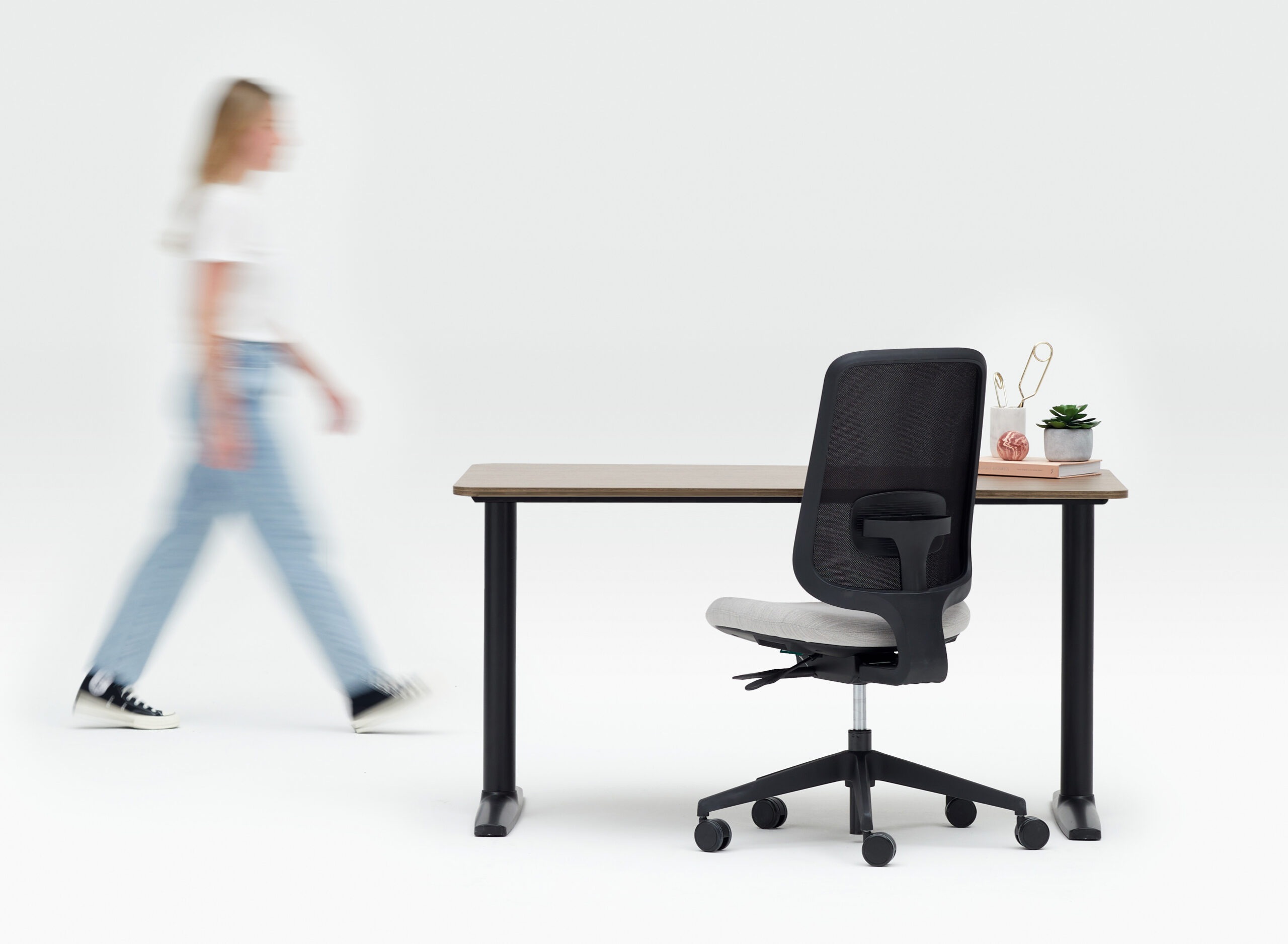 Person Walking Towards a Fixed Height Desk and a Black Frame Office Chair With No Arms